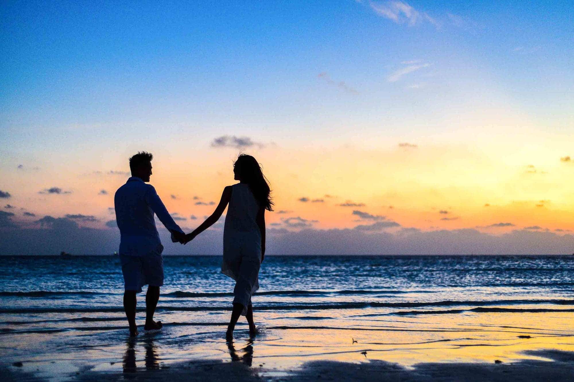 A couple holding hands, walking along the shoreline at sunset.